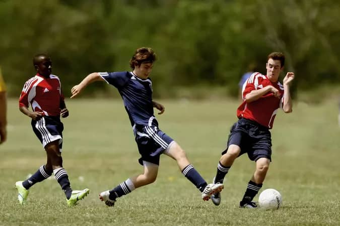 Three young soccer players are engaged in a match on a grassy field, illustrating the intensity and fairness promoted by the mercy rule in football, with one player dribbling the ball while two others defend.
